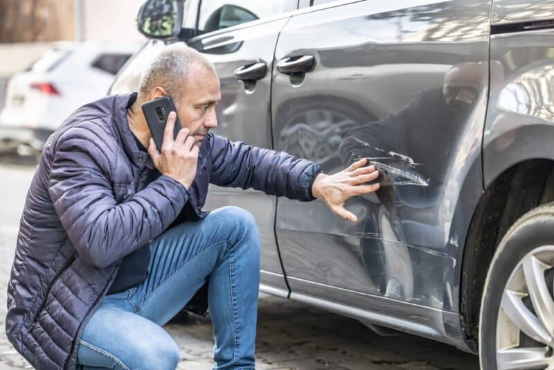 A man examines damage to his car while on the phone to his car insurance agency