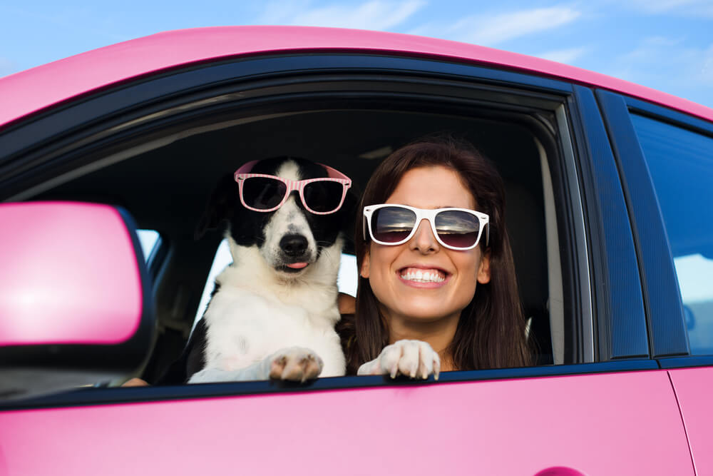 Woman and dog wearing sunglasses smiling from the window of their pink car.