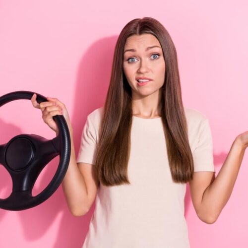 Pretty girl with pink background holding a steering wheel and shrugging.