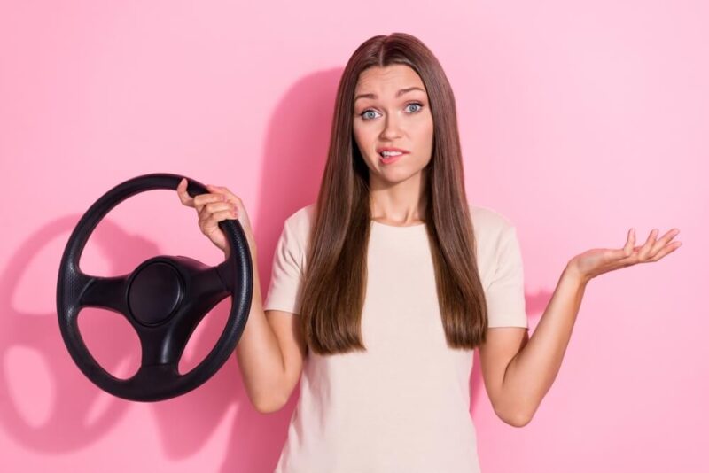 Pretty girl with pink background holding a steering wheel and shrugging.